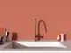 Küchenarmatur in glänzendem Rosé mit schlankem Auslauf und moderner Form. Ideal Standard, platziert über einem weißen Spülbecken vor einer rötlichen Wand.